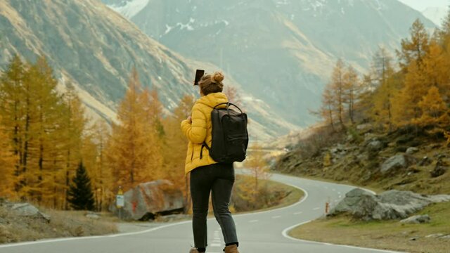 Woman traveller on hike in epic autumn mountains. Happy and excited young woman in outdoor adventure gear outfit walk on beautiful mountain road, spin around, record memories and experiences on phone