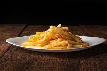 French fries placed on a white plate on rustic wood with black background, selective focus.
