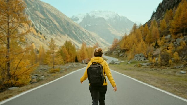 Camera follows young female traveller in yellow puffy jacket and backpack, walk in middle of epic empty road in mountains, surrounded by nature, autumn or fall colours. Travel road trip wanderlust