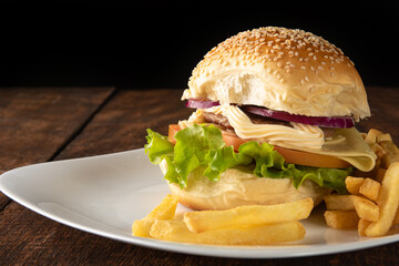 Hamburger and French fries placed on a white plate on rustic wood with black background, selective focus.