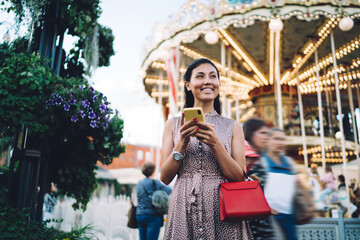 Charming hipster girl with cellphone device enjoying leisure day for recreating in Amusement Park, cheerful solo traveller with smartphone gadget smiling in urban city feeling pleasure outdoors