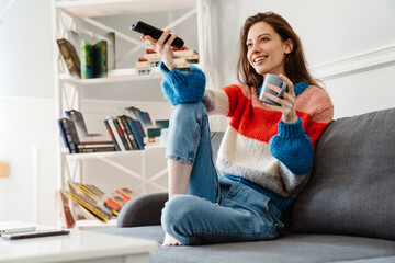 Photo of girl drinking coffee and using remote control while watching tv - Powered by Adobe