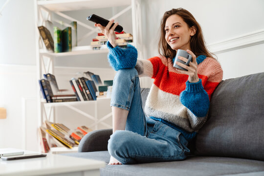 Photo of girl drinking coffee and using remote control while watching tv