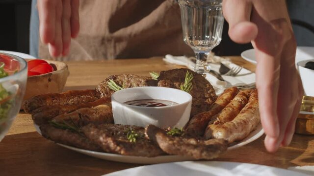 Close up of unrecognizable cook is putting big plate with dish of several types of meat and tomato sauce in the middle of it on the table