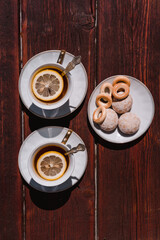 Beautiful white ceramic tableware for tea drinking. Two white cups of black tea with lemon on a wooden table, and delicious bagels and gingerbread in a saucer.