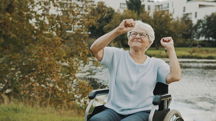 Happy old woman, grandmother, dancing with hands in the wheelchair near the river . High quality photo