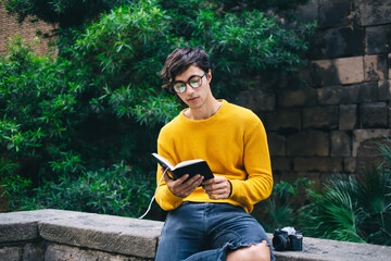 Obraz premium Serious concentrated handsome male reader in eyewear for vision correction enjoying free time on holly and self education, thoughtful hipster guy holding book with interesting plot resting outdoors