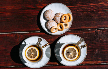Beautiful white ceramic tableware for tea drinking. Two white cups of black tea with lemon on a wooden table, and delicious bagels and gingerbread in a saucer.