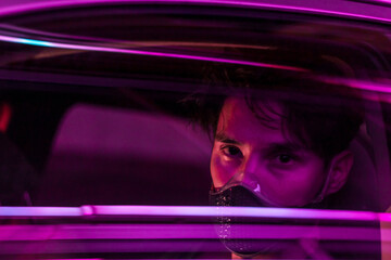 a young man in a face mask driving a car in the night city with neon lights reflecting from the glass of his car