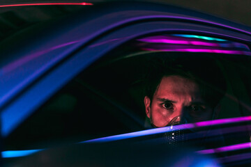 a young man in a face mask driving a car in the night city with neon lights reflecting from the glass of his car