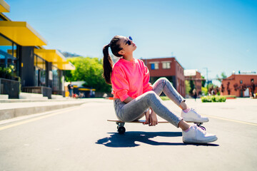 Attractive female teenager in stylish sunglasses resting at longboard during sport hobby in city,...