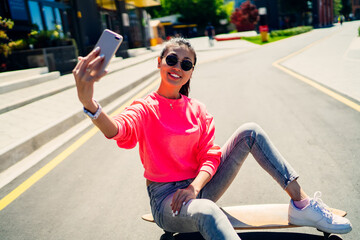 Joyful skater dressed in fashionable streetwear clicking content selfie during leisure in city, sincerely hipster girl sitting at longboard and creating influence vlog for sharing to social networks