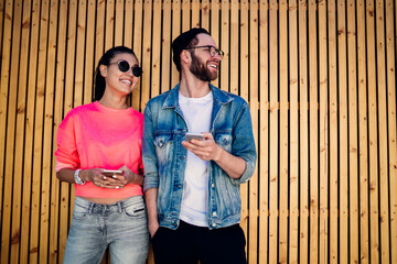 Happy male and female influence bloggers standing near promotional background and smiling, sincerely hipster guys using cellphone gadgets for network browsing on leisure, concept of pda technology