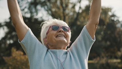 Cool grandmother with pink sunglasses and headphones stretching in the park. Recreation in old...