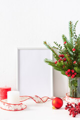 Christmas poster mock up with vertical frame, fir tree branches and hawthorn twigs in a vase and red balls with candles on white wall background, new year and christmas concept