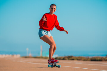 Happy smiling carefree woman in red hoodie and jeans shorts riding surf skateboard and showing shaka sign. Summer fun outdoor activities concept. Street culture.