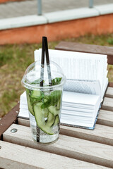 Cucumber lemonade stands on street against background of book. Tasty summer green lemonade with straw. Vertical photo.