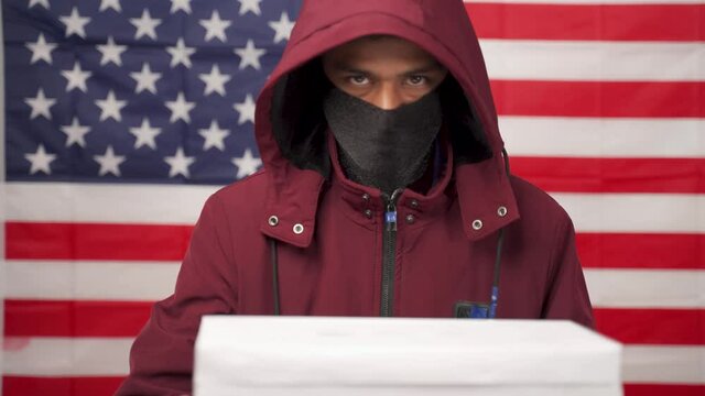 Man in hoodie with face covered casting Vote at polling booth with US falg as background - Concept of unknown voting or vote rigging in US elections.