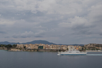 Fototapeta na wymiar A white ferry boat traveling past the city of Corfu, Greece on a cloudy summer day. View from the sea.