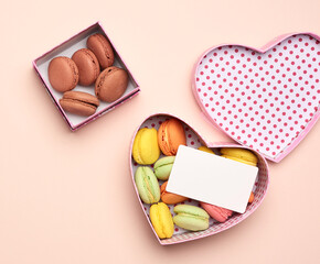round baked multicolored macarons lie in a pink cardboard box in the shape of a heart on a beige background