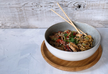 Udon noodle with fried chicken and vegetables in a grey bowl with sticks on the bamboo serve plate and wood background