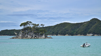 Naklejka premium Timbered and rocky desert island and white sailboat against a background of thick forest in Torrent Bay lagoon, Abel Tasman National Park, South Island, New Zealand