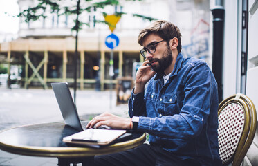 Serious male freelancer talking on smartphone and typing on laptop