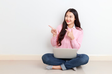 Young attractive Asian woman sitting with laptop on her legs finger pointing up showing copy space with smiley face.