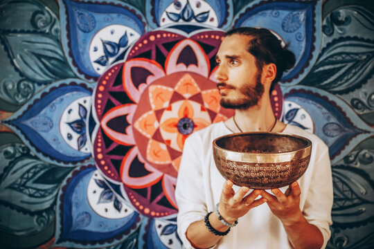 stylish bearded man holding a large Tibetan bowl on the background of a large color picture of a mandala