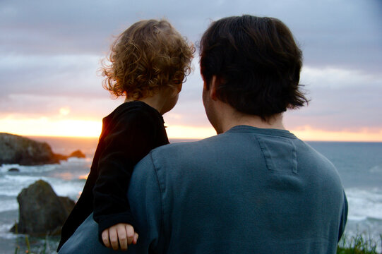 Back view of a father holding his toddler girl as they look at the sunset over a rocky Pacific Northwest (USA) beach