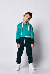 Little brunette kid in colorful tracksuit and sneakers on a gray background.