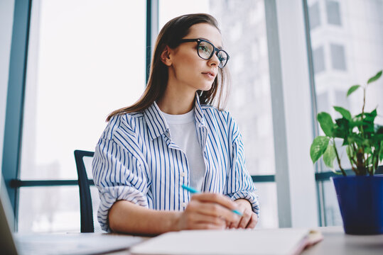 Thoughtful female writer in smart casual clothing sitting at table desktop with cropped laptop and working with ideas for SEO text, pensive content manager in spectacles planning project organisation