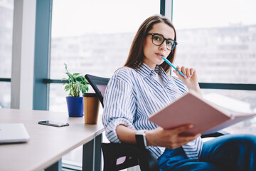 Portrait of thoughtful female writer in spectacles for eyes correction sitting at desktop holding personal notebook, pensive woman looking at camera working on creation project in office interior