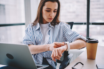 Caucasian woman in smart casual wear checking time to deadline of startup project using wearable smartwatch in modern workspace, skilled woman with modern laptop computer touching clock display