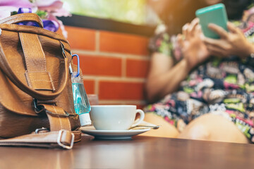 Mini alcohol gel bottle to kill Corona Virus(Covid-19) hang on a brown leather shoulder bag with hot coffee on table in cafe.New normal lifestyle.Health care concept.Selective focus on alcohol gel