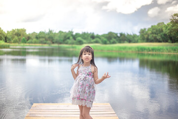 Close-up view of a young Asian girl, lovely, cute wearing a colorful dress, sitting by a pond in the village, closely supervised during school holidays