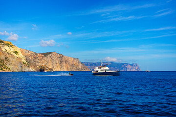 Powerboat and hydrocycle in the sea against high coast and blue sky with spindrift clouds in Crimea. Seascape. Local tourism. Black sea