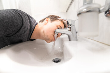 young man drink tap water by mouth out of sink in bathroom with lips and tongue in flowing water...