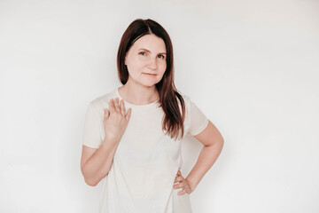 Beautiful brunette woman keeps hands on cheek, dressed in casual white t shirt, has minimal makeup, poses indoor on a white background. Copy, empty space for text