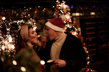 Couple in love relationship believe in meracle, kissing against the chrismas decoration outdoor in the eve, look at each other and drink the warm drinks