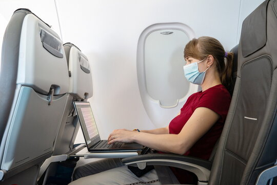 Caucasian young woman wearing pandemic medical face mask working on her laptop on an airplane seat during flight