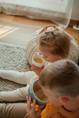 Little brother and sister in cozy knitted sweaters are drinking tea at home. Portrait of cute little brother and sister. Fall.