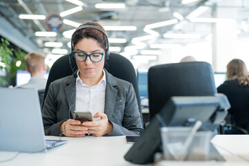 Business woman dressed in a headset is bored and uses a smartphone while sitting at a desk. Female...