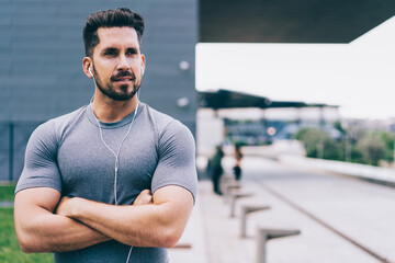 Handsome male runner dressed in active wear and electronic earphones pondering on healthy workout,Caucasian muscular trainer with crossed hands using headphones for listening motivation music playlist