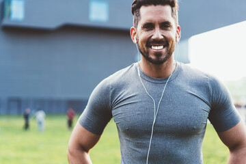 Half length portrait of cheerful Caucasian trainer with perfect smile and muscular body posing during morning workout, happy male bodybuilder in electronic headphones listening positive music