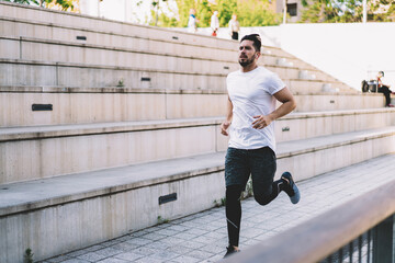 Obraz premium Strength Caucasian male in active wear jogging at urban city street, sportsman in earphones listening playlist songs while doing cardio training keeping healthy lifestyle and perfect body shape