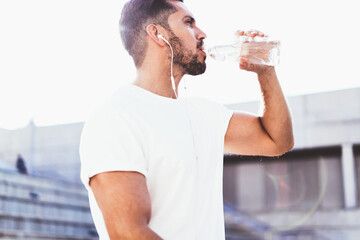 Fototapeta na wymiar Handsome male athlete 30s drinking water after cardio jogging workout listening music via earphones equipment, Caucasian sportsman in activewear feeling thirsty on physical strength training