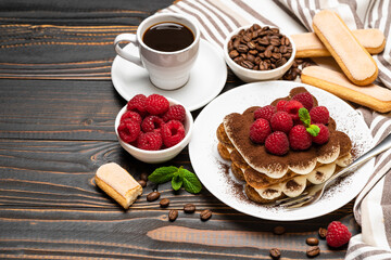 portion of Classic tiramisu dessert with raspberries, savoiardi cookies and cup of espresso coffee isolated on wooden background