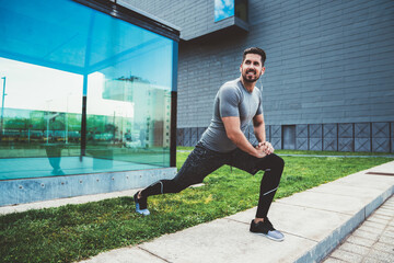 Caucasian sportsman in active wear and earphones doing exercises warm up for training outdoors, experienced male athlete preparing for jogging keeping healthy lifestyle and physical strength