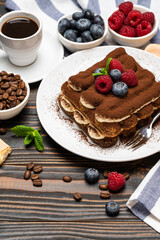 portion of Classic tiramisu dessert with raspberries and blueberries, savoiardi cookies and cup of espresso coffee isolated on wooden background